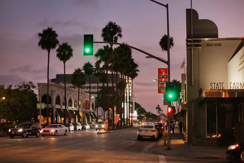 View of Culver City street with stoplights, white cars, and palm trees sillhouetted against a purple sunset sky